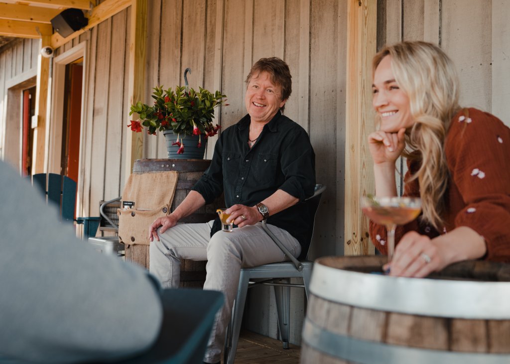 Three people are enjoying a casual outdoor gathering, seated on a wooden deck with a rustic barrel and potted plants nearby in Williamson County, Tennessee.