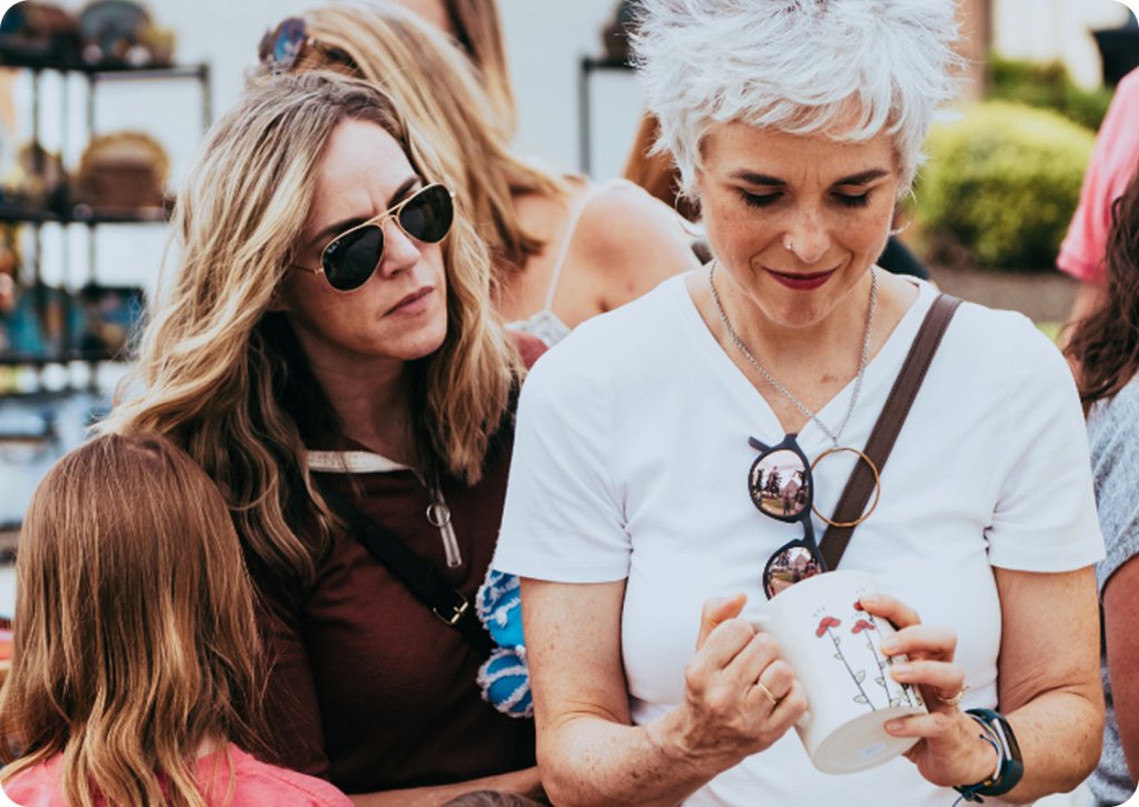 Two women, one with long blonde hair and the other with short gray hair, are engaged in conversation at an outdoor event, with one holding a white mug with a floral design in Williamson County, Tennessee.