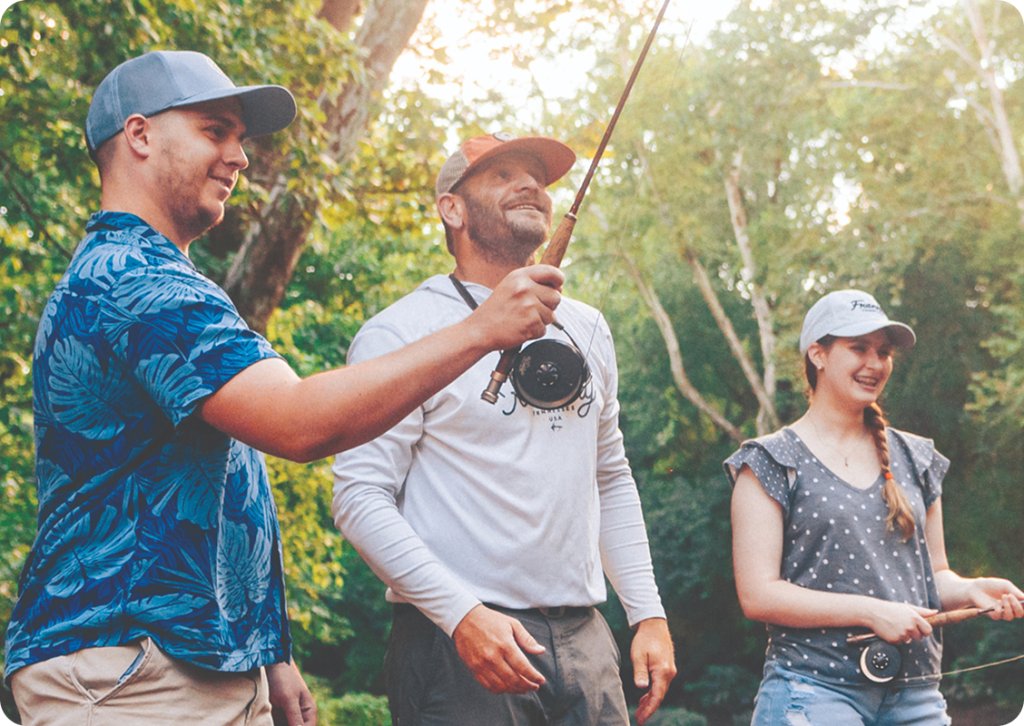 Three friends, dressed in casual outdoor attire, enjoy a sunny day fishing in a lush, green forest setting in Williamson County, Tennessee.
