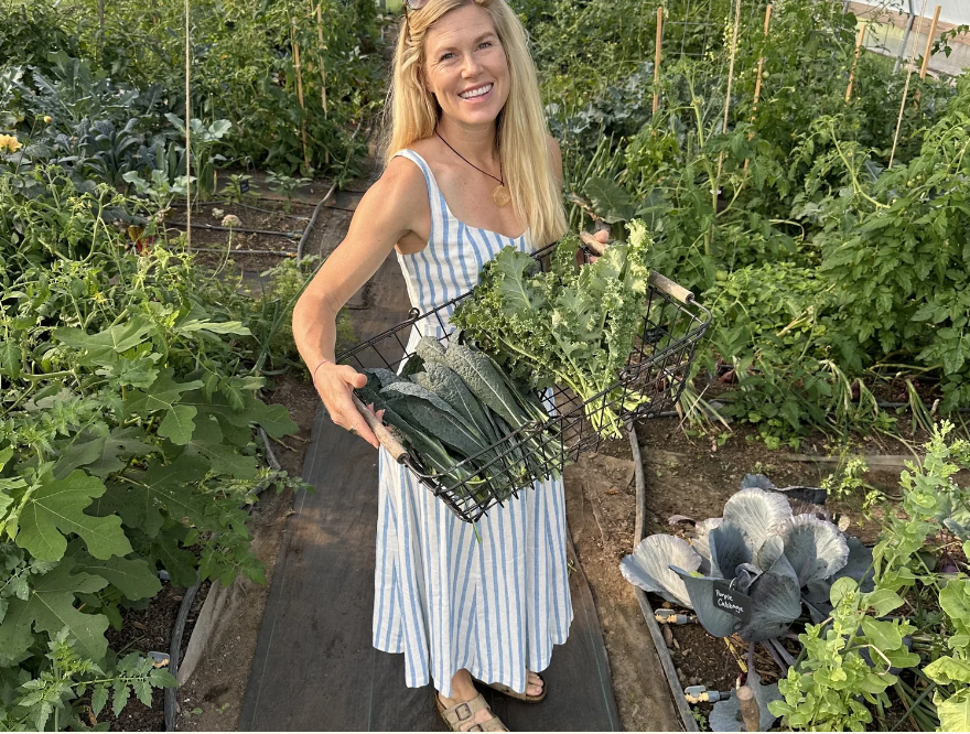 A woman in a blue and white striped dress smiles while holding a basket of fresh kale and other leafy greens in a lush garden in Williamson County, Tennessee.