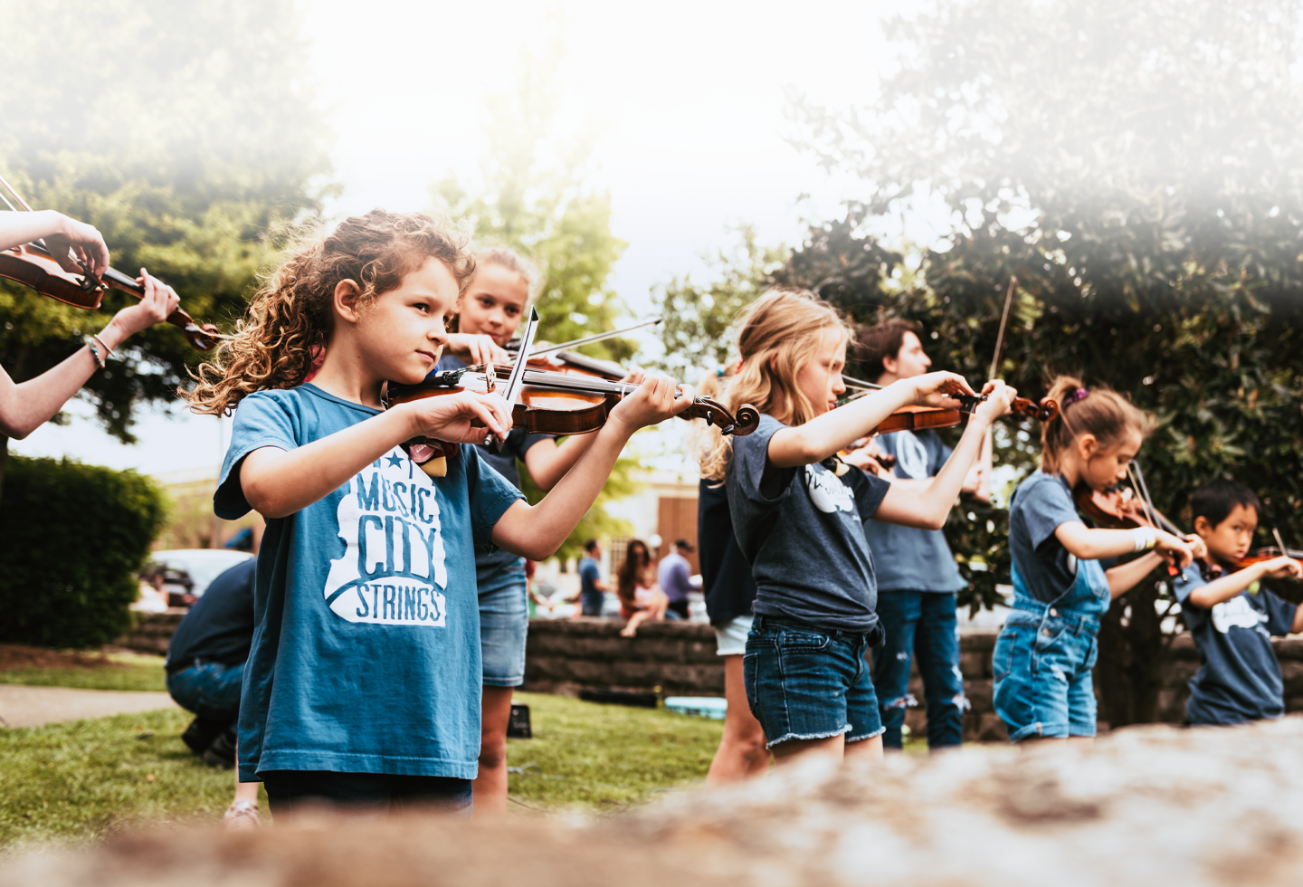 A group of young violinists, dressed in blue shirts, perform together in an outdoor setting surrounded by trees and greenery in Williamson County, Tennessee.