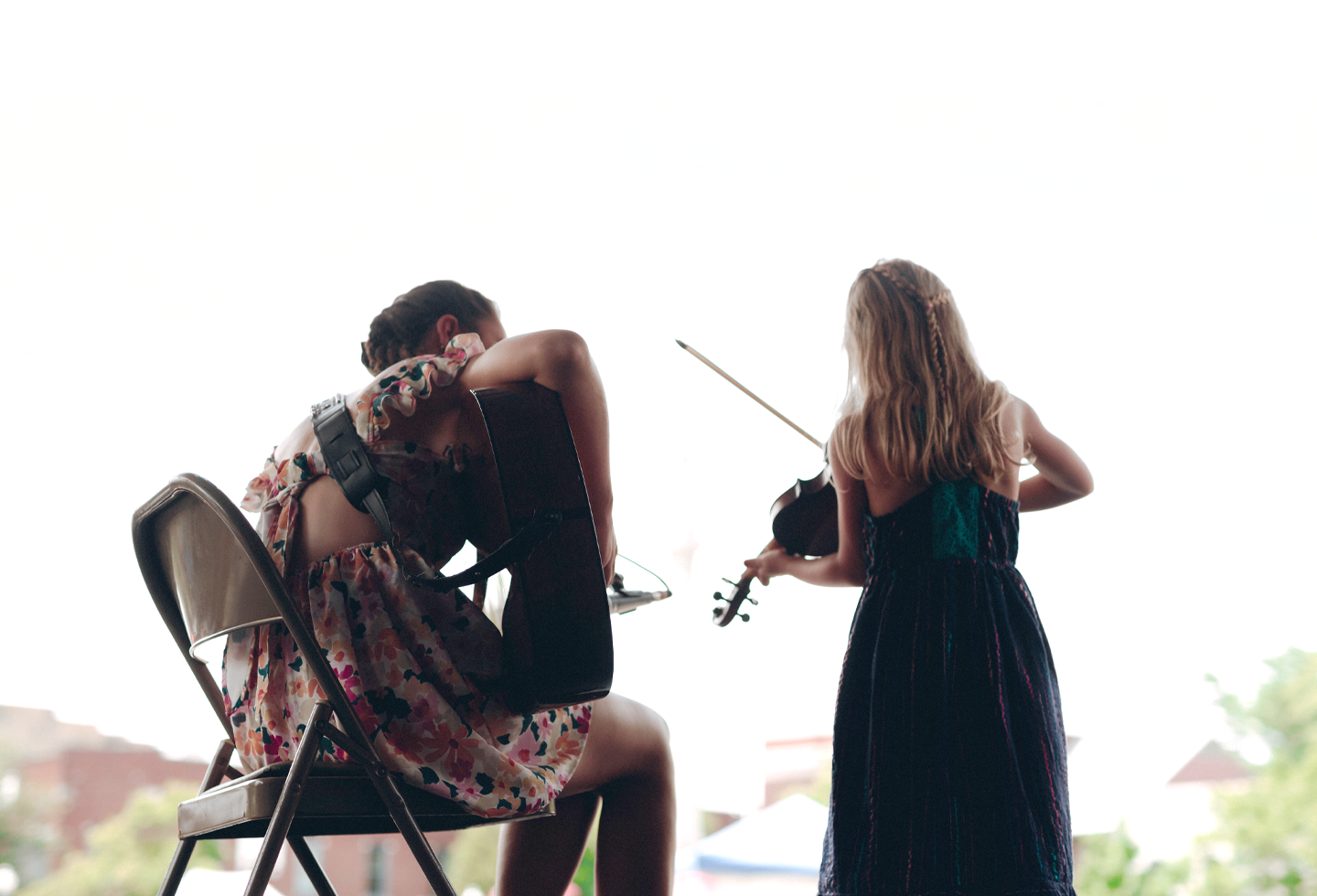 Two young girls, one playing guitar and the other violin, are seated outdoors, facing away from the camera in Williamson County, Tennessee.