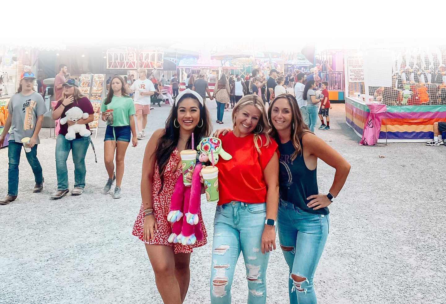 Three women in summer outfits pose for a photo at a lively outdoor fair, surrounded by colorful rides and games in Williamson County, Tennessee.