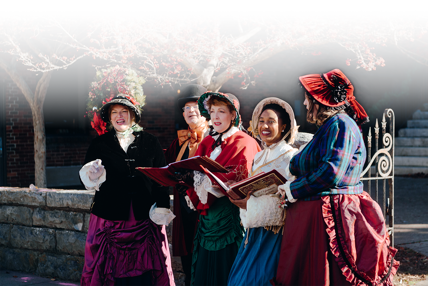 A group of five women dressed in vibrant Victorian-era costumes sing joyfully in front of a brick building adorned with a festive wreath in Williamson County, Tennessee.