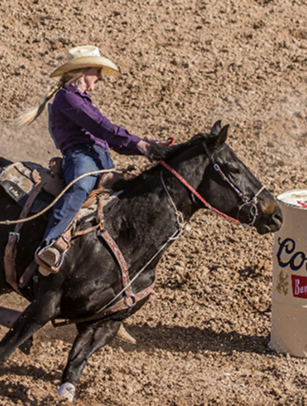 A determined cowgirl in a purple shirt and blue jeans rides a black horse around a barrel, kicking up dust in a competitive barrel racing event in Williamson County, Tennessee.