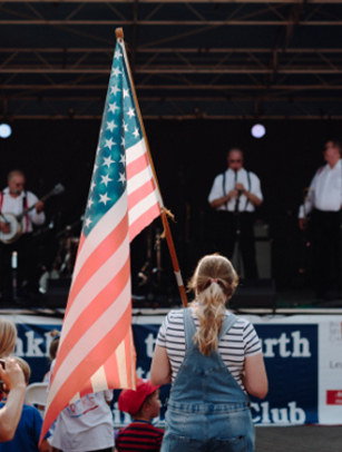 A woman in overalls holds an American flag while a band performs on stage at a patriotic event in Williamson County, Tennessee.