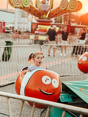 A young girl with pigtails rides a vibrant orange clownfish-themed bumper car at a bustling fairground, her face lit up with pure joy as she navigates the colorful amusement park rides in Williamson County, Tennessee.