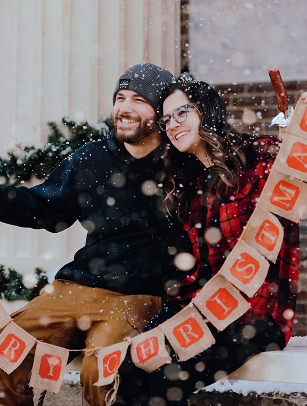 A couple dressed in winter attire, including a red and black plaid shirt and a gray beanie, are joyfully posing together in the snow, holding a banner that reads 'Merry Christmas' in Williamson County, Tennessee.
