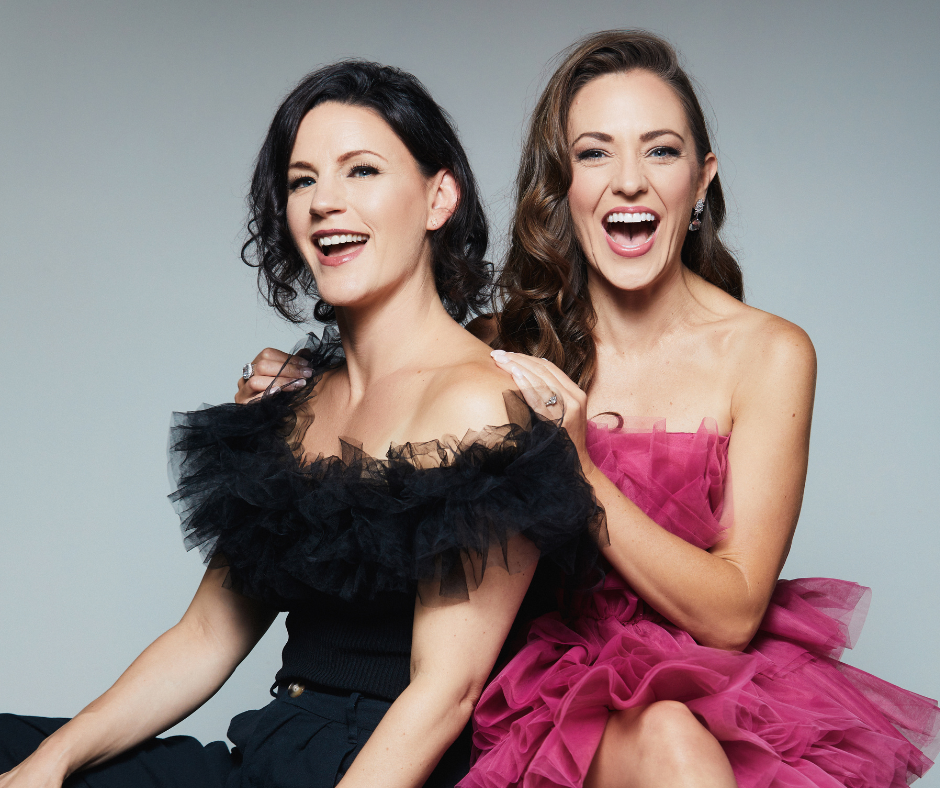 Two women in elegant dresses laugh joyfully, one in a black off-the-shoulder top with a ruffled tulle overlay and the other in a vibrant pink strapless tulle dress in Williamson County, Tennessee.