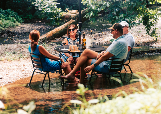 Four people enjoy a casual outdoor gathering at a small table set in a shallow stream, surrounded by lush greenery in Williamson County, Tennessee.