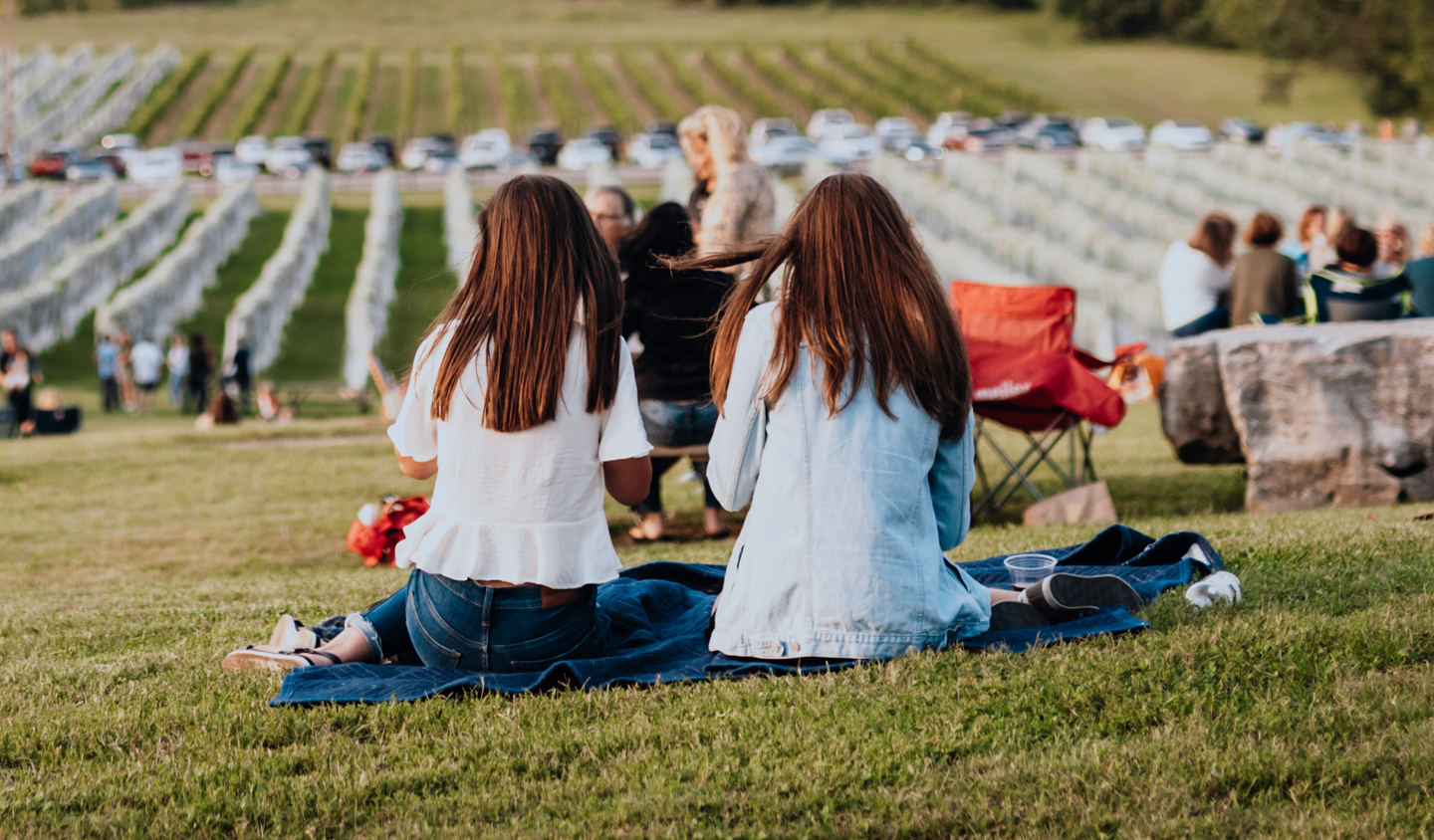 Two women with long hair sit on a blue blanket in a grassy field, enjoying a sunny day with a view of a vineyard and parked cars in the distance in Williamson County, Tennessee.
