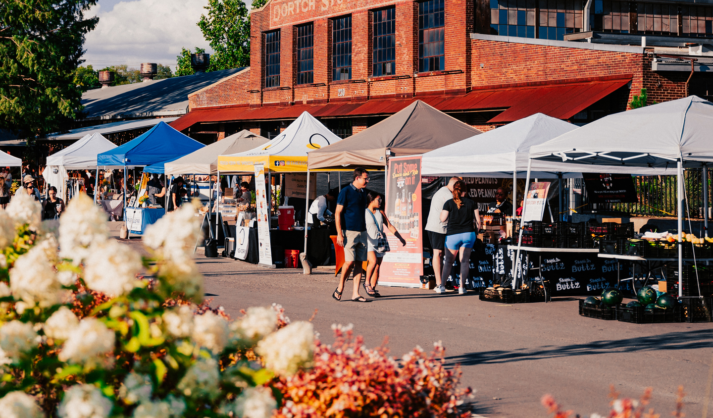 A bustling outdoor market with colorful tents and a historic brick building in the background, surrounded by vibrant flowers and trees in Williamson County, Tennessee.