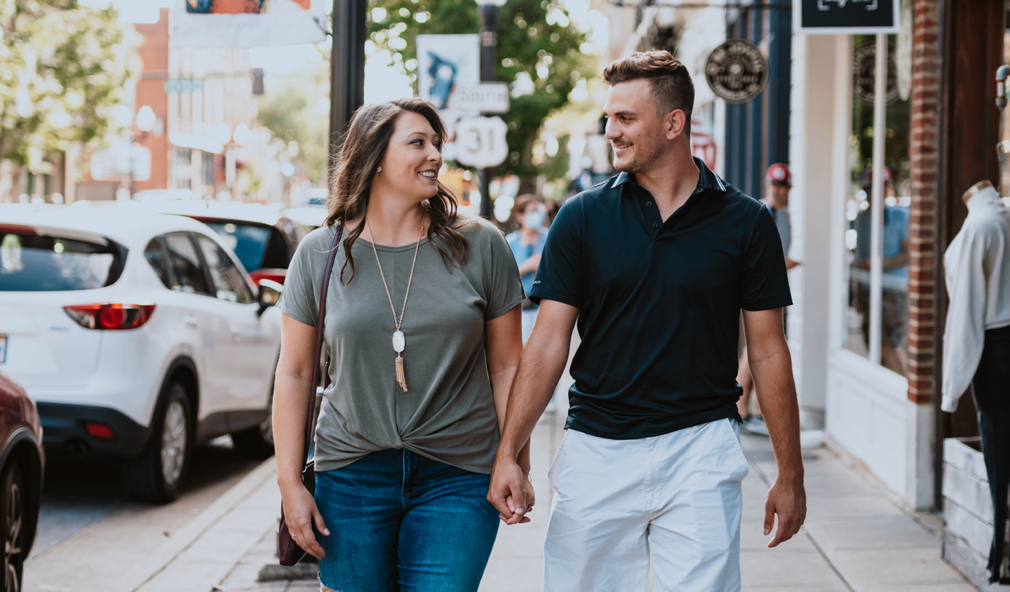A couple walks hand in hand down a bustling city street, surrounded by parked cars and storefronts in Williamson County, Tennessee.