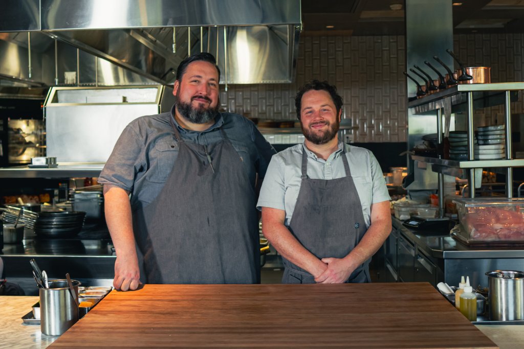 Two chefs stand side by side in a professional kitchen, both wearing aprons and smiling warmly at the camera in Williamson County, Tennessee.