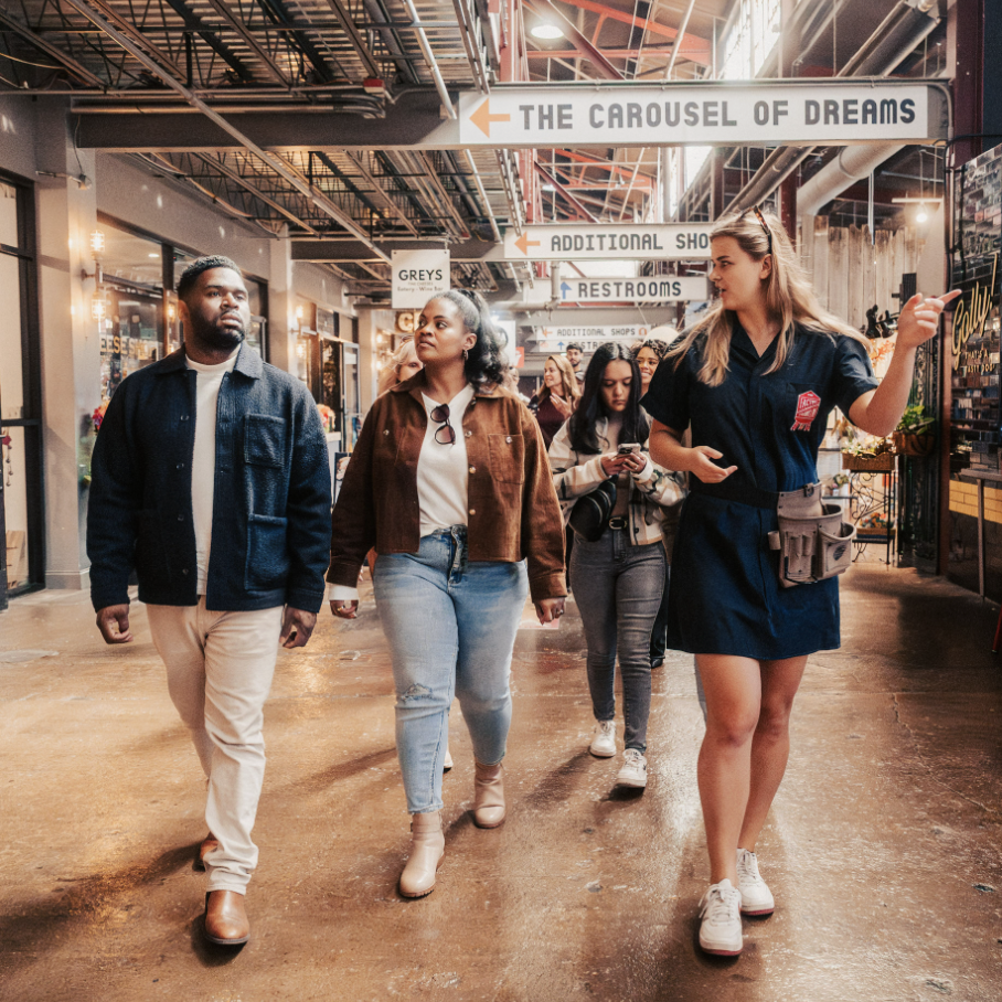 A diverse group of people walks through a bustling indoor market with high ceilings and hanging signs in Williamson County, Tennessee.