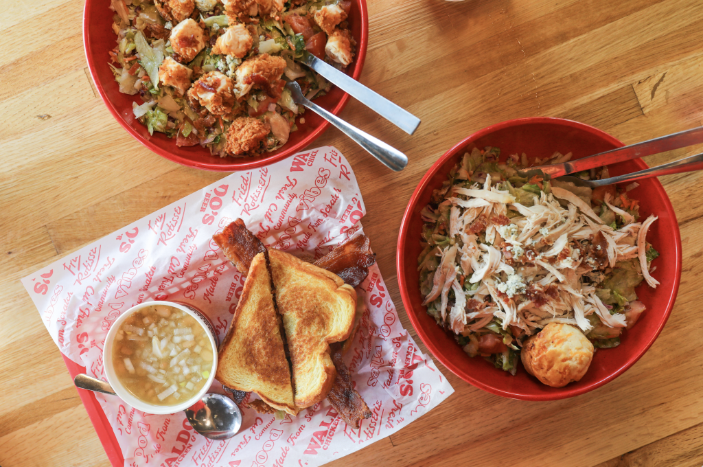 A wooden table holds three red bowls filled with crispy fried chicken salad, a grilled cheese sandwich with bacon, and a bowl of creamy chicken soup, all served on branded paper with utensils in Williamson County, Tennessee.
