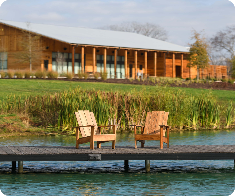 Two wooden Adirondack chairs sit invitingly on a dock extending into a serene pond, with a modern wooden building in the background in Williamson County, Tennessee.