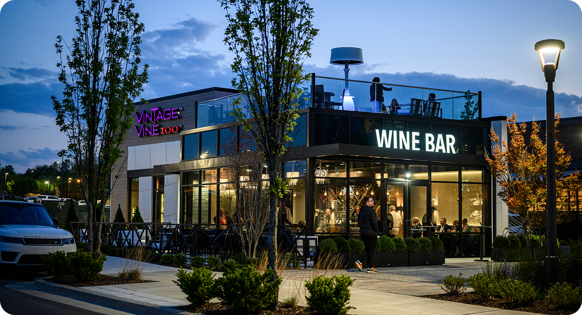 A modern wine bar with a rooftop patio, illuminated by a purple neon sign and outdoor lighting, surrounded by trees and a parking lot at dusk in Williamson County, Tennessee.