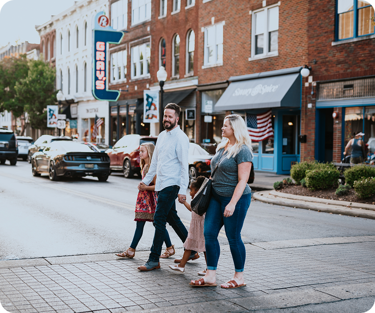 A family of four, including two adults and two children, walks hand in hand down a bustling city street lined with shops and cars in Williamson County, Tennessee.