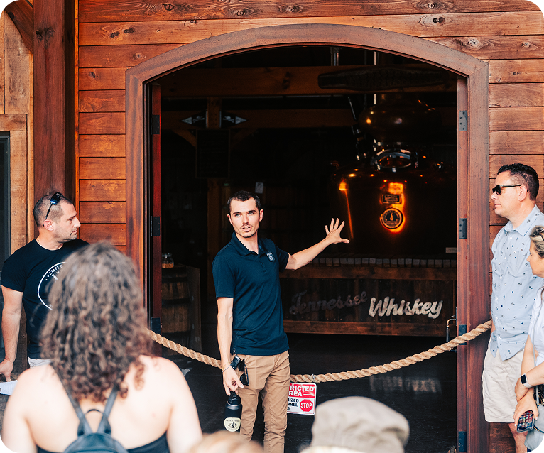 A group of people gather around a wooden doorway, where a man gestures towards a large copper still inside a dimly lit whiskey distillery in Williamson County, Tennessee.