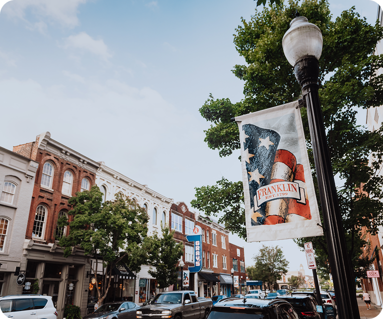 A quaint town street lined with historic buildings, trees, and an American flag banner hanging from a lamppost in Williamson County, Tennessee.