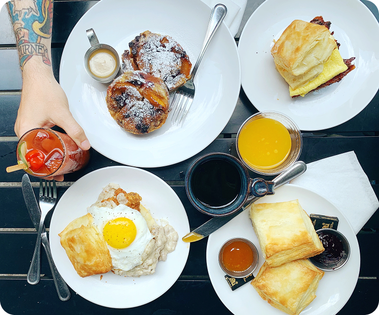 A hand holding a glass of red drink with a straw, next to a table set with plates of pastries, biscuits, eggs, and various condiments in Williamson County, Tennessee.