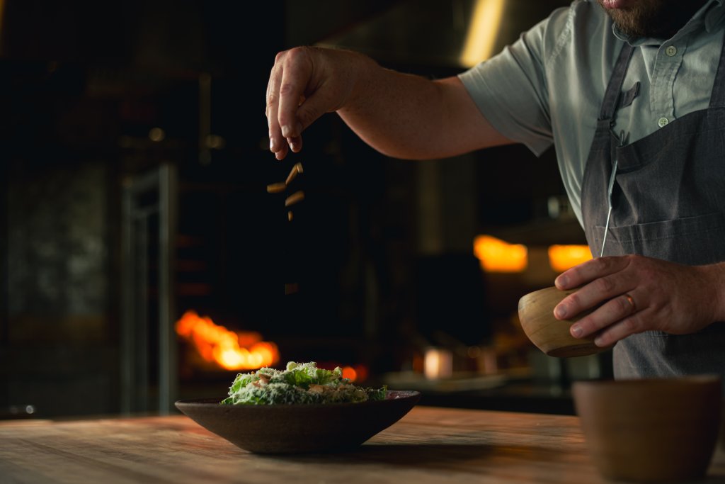 A chef sprinkles seasoning over a dish of greens in a rustic kitchen with a warm fire in the background in Williamson County, Tennessee.