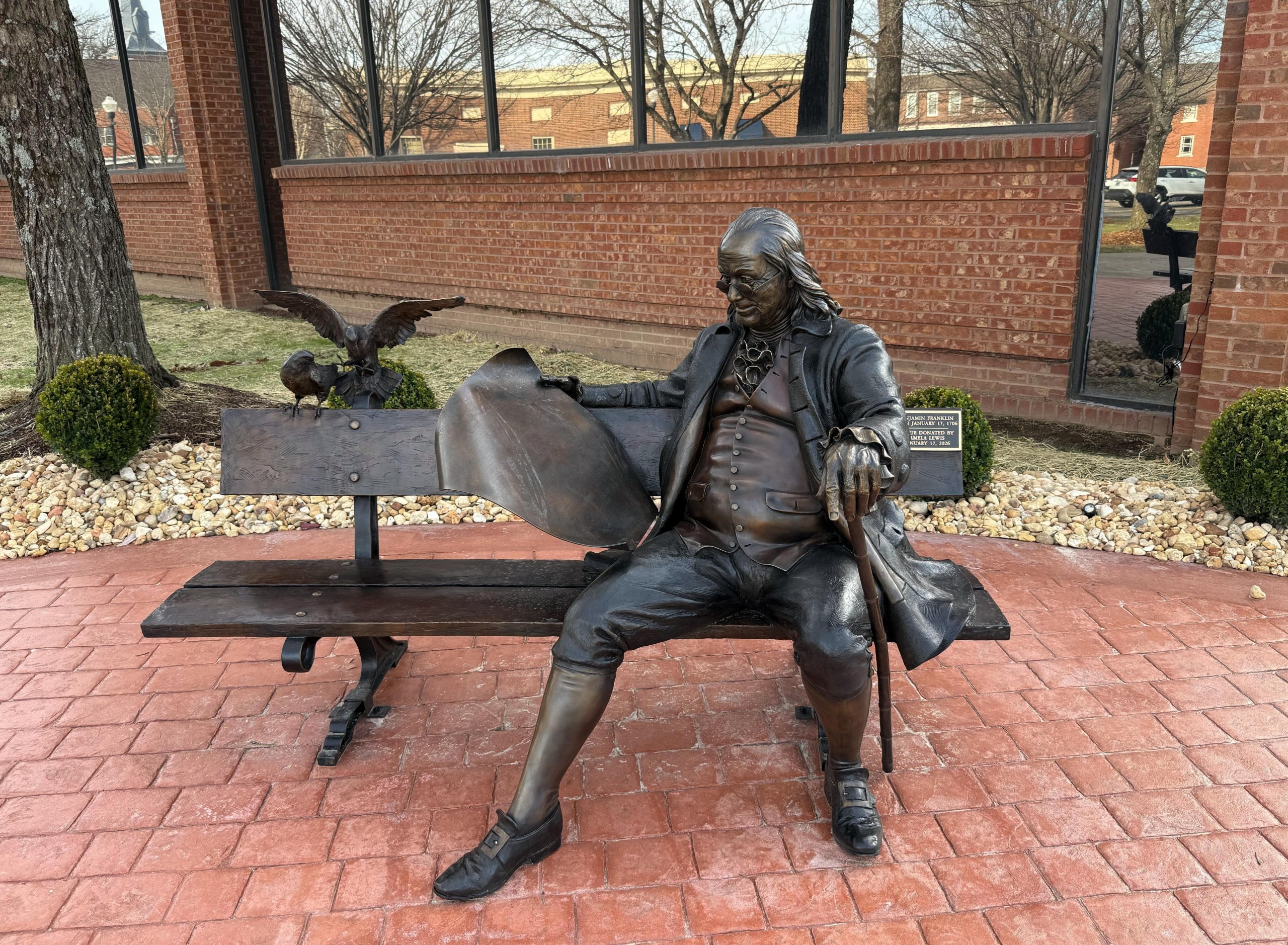 What's New & Coming A bronze statue of Benjamin Franklin seated on a bench, holding a book and accompanied by an eagle and a turtle, set against a backdrop of a brick building and landscaped garden in Williamson County, Tennessee.