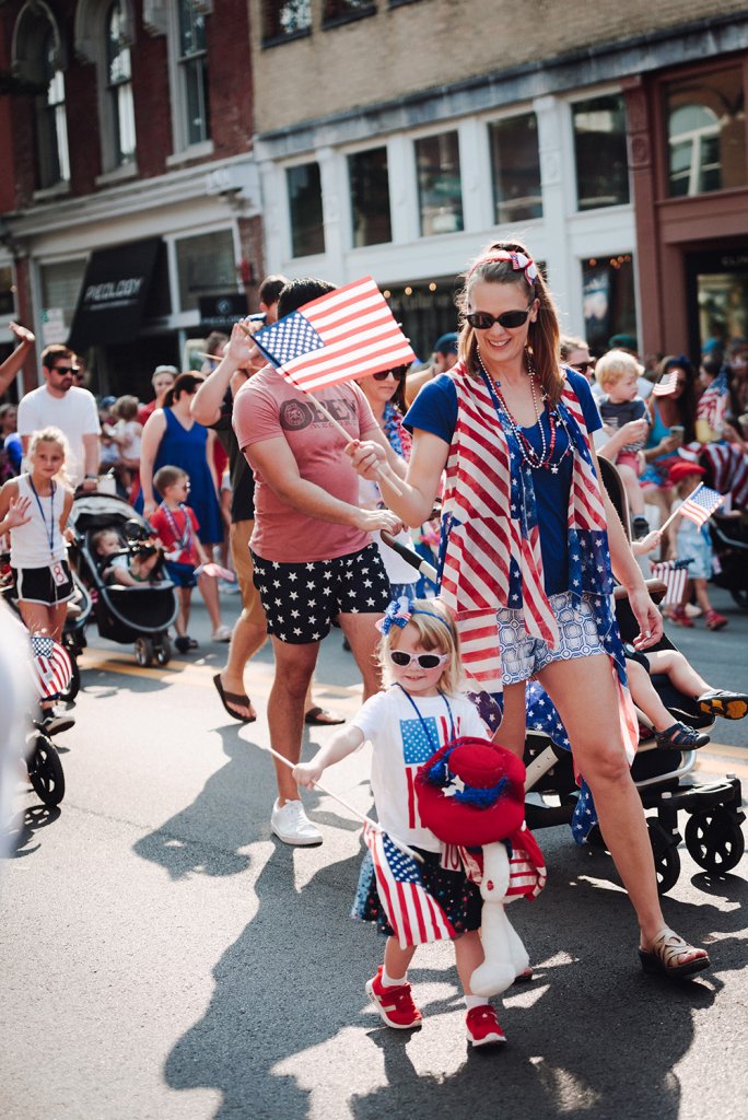 A family dressed in patriotic attire waves American flags while marching in a Fourth of July parade, surrounded by a crowd of spectators in Williamson County, Tennessee.