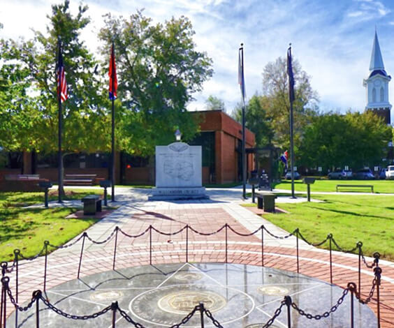 A serene park features a central monument surrounded by flags, benches, and lush greenery in Williamson County, Tennessee.