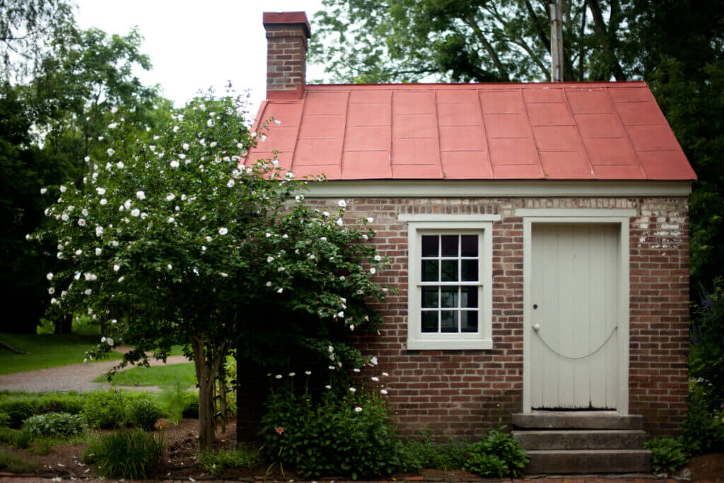 A quaint brick cottage with a red metal roof is nestled among lush greenery, featuring a white-framed window and a charming white door with a small window pane in Williamson County, Tennessee.