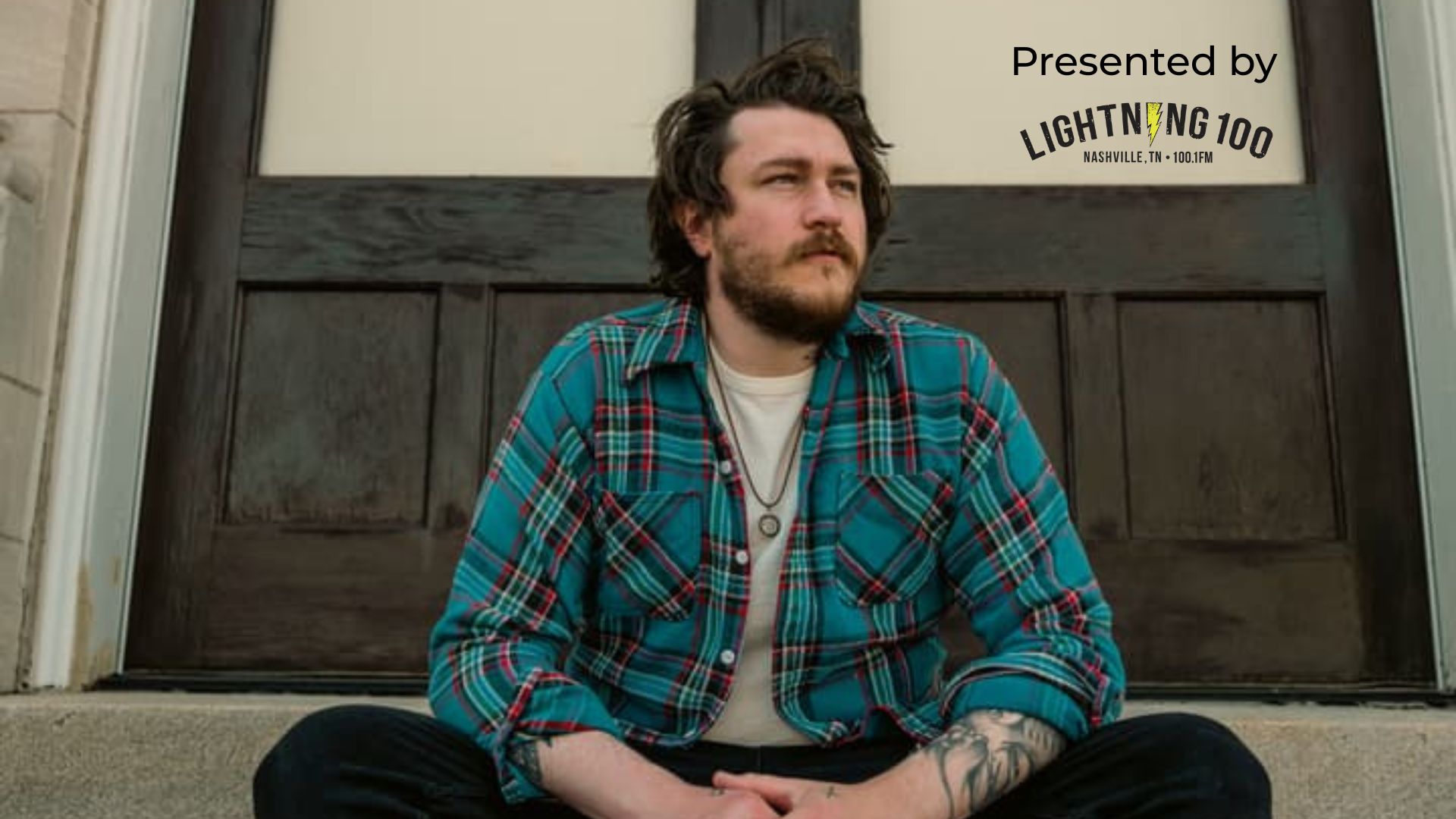 A man with a beard sits cross-legged on steps, wearing a blue plaid shirt and a necklace, with a wooden door behind him in Williamson County, Tennessee.