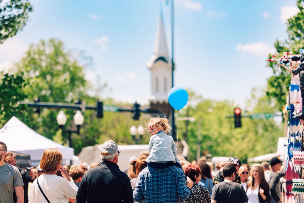 A lively outdoor festival scene with a crowd of people, a child holding a blue balloon, and a church steeple in the background in Williamson County, Tennessee.
