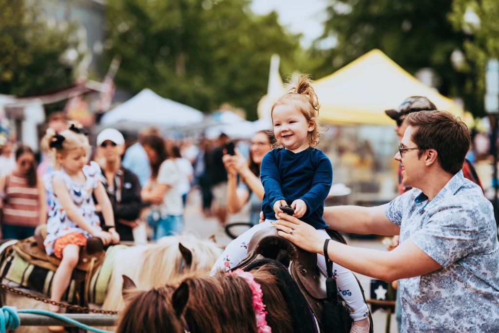A young girl in a blue shirt rides a pony at a lively outdoor festival, smiling broadly as her father guides the horse in Williamson County, Tennessee.