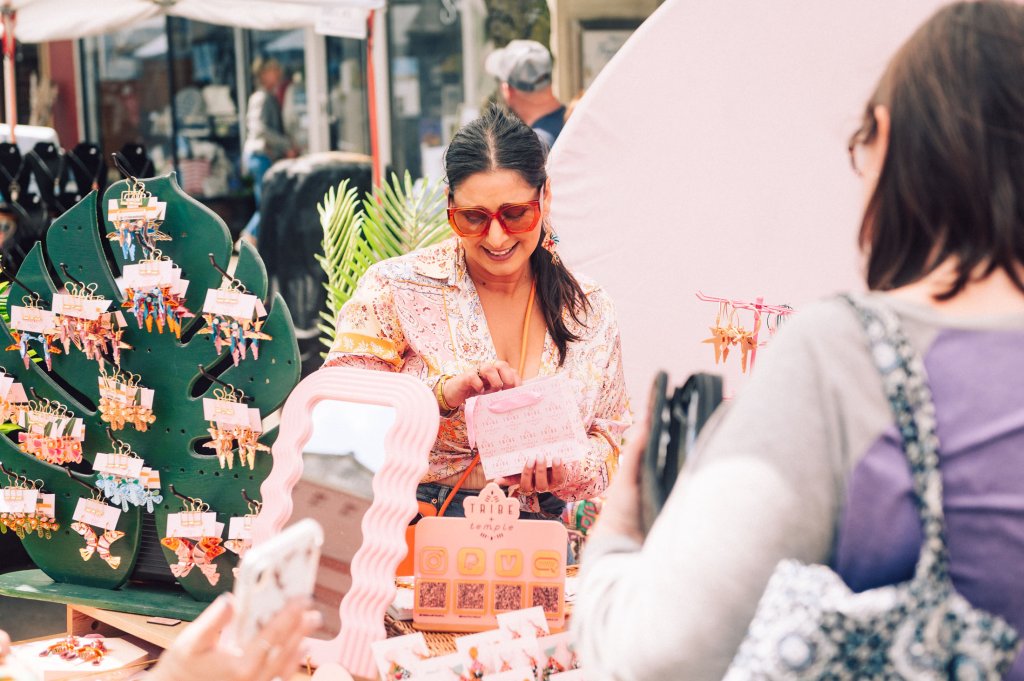 A woman in a colorful blouse and sunglasses is selling handmade jewelry at an outdoor market, surrounded by vibrant earrings and accessories in Williamson County, Tennessee.