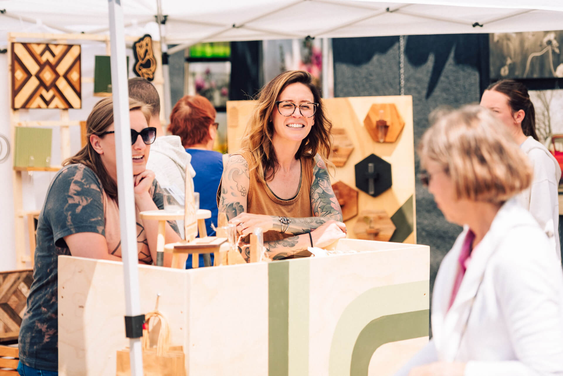 A group of women are gathered around a wooden booth at an outdoor market, browsing and chatting about the handcrafted items on display in Williamson County, Tennessee.