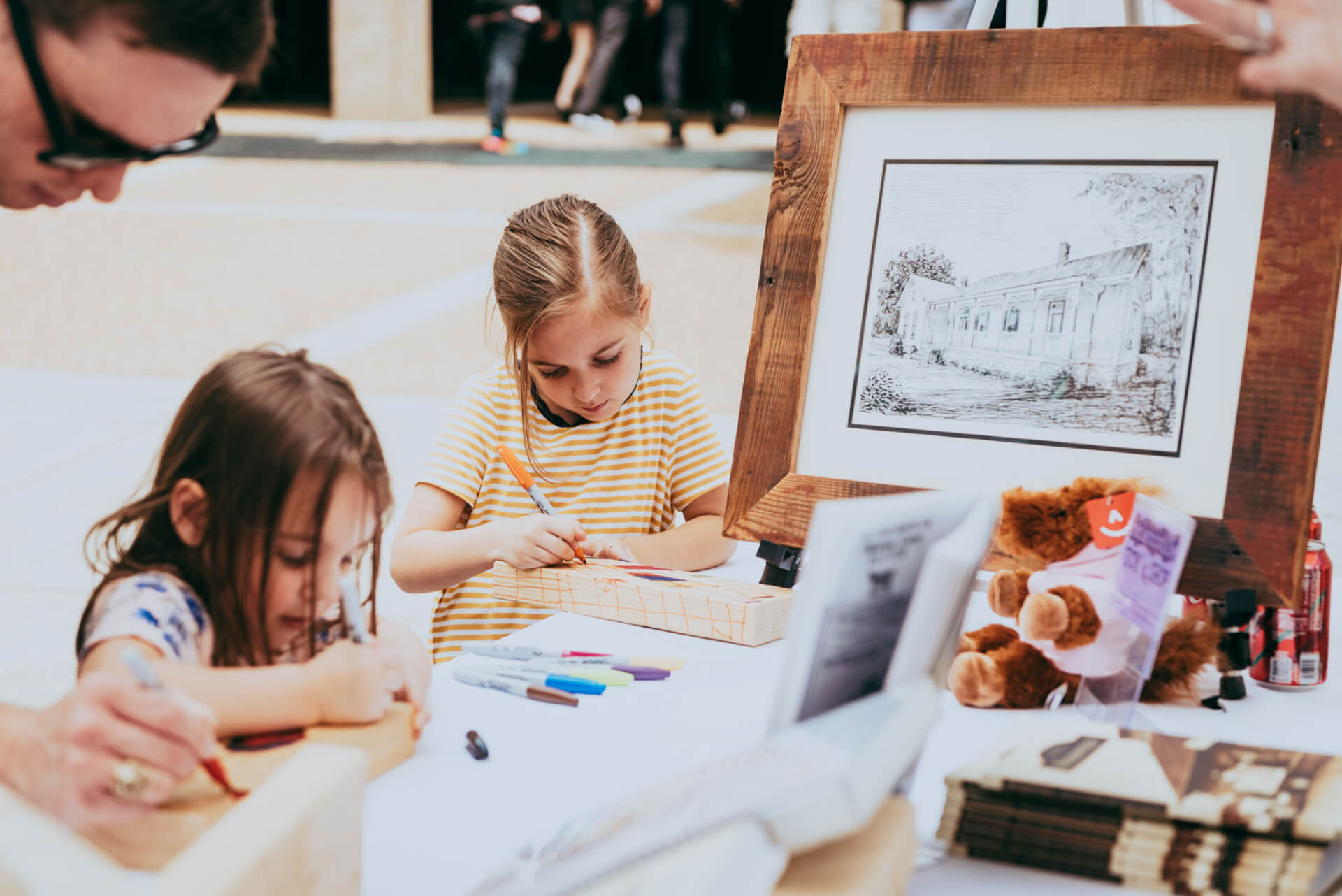 Two young girls are engrossed in drawing at a table, surrounded by art supplies and a framed picture of a house, while an adult assists them in Williamson County, Tennessee.