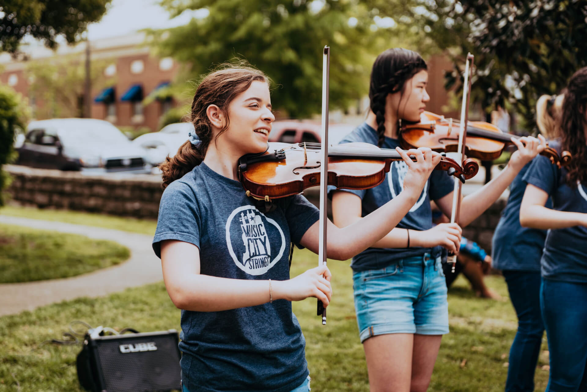 A group of young women, dressed in matching blue shirts, play violins together in a park setting, with trees and a building in the background in Williamson County, Tennessee.