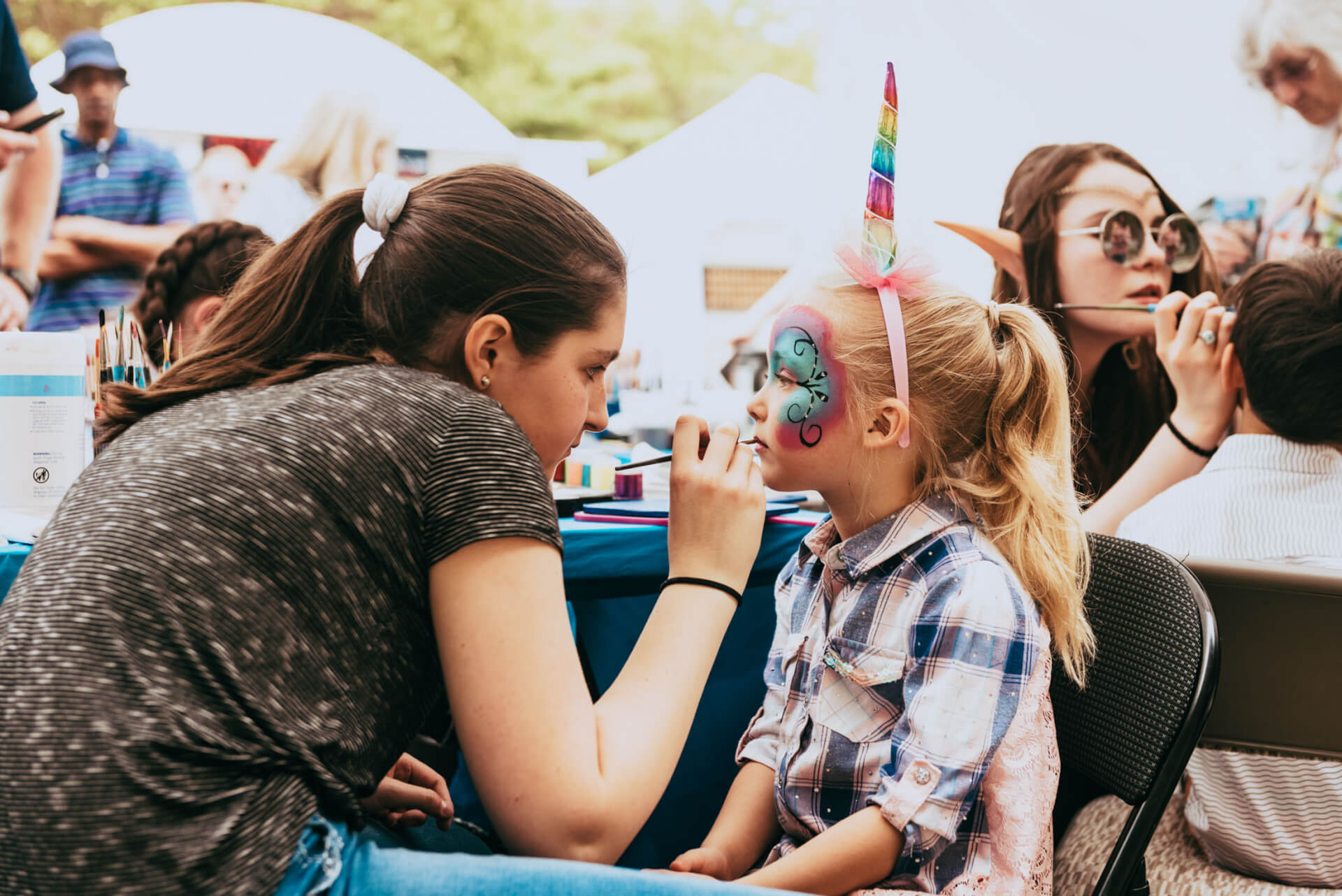 A woman carefully paints a young girl's face with intricate designs at a face painting booth, while the girl wears a colorful unicorn horn headband in Williamson County, Tennessee.