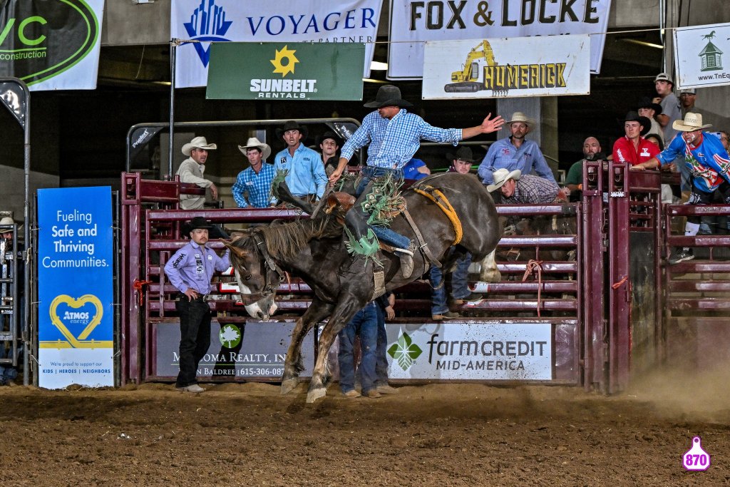 A cowboy in a blue checkered shirt and cowboy hat is riding a bucking bronco in a rodeo arena, with spectators and event sponsors visible in the background in Williamson County, Tennessee.