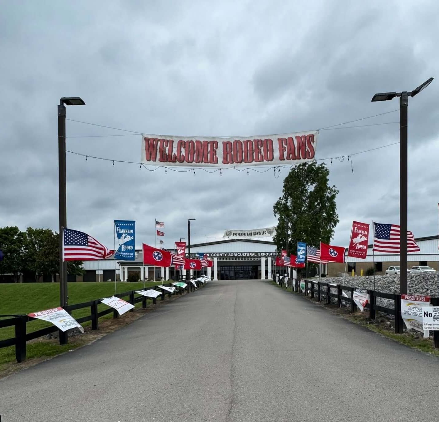 A wide, empty road leads towards a large building adorned with numerous flags, including American and rodeo-themed banners, under a cloudy sky in Williamson County, Tennessee.