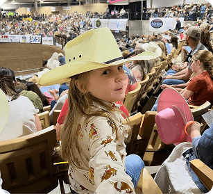 A young girl in a cowboy hat smiles at the camera amidst a crowded rodeo arena in Williamson County, Tennessee.