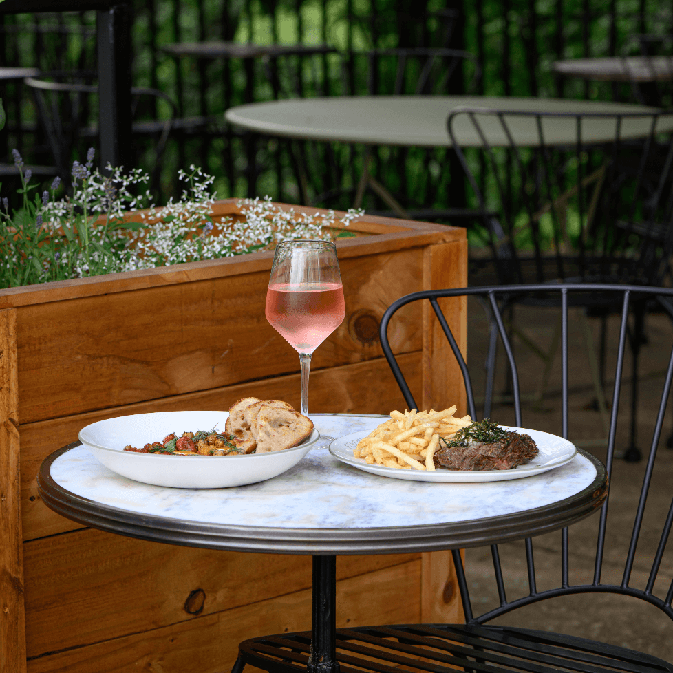 A charming outdoor dining setup features a small round table with a marble top, adorned with a glass of rosé wine, a plate of steak and fries, and a bowl of salad, all set against a backdrop of lush greenery and wooden planters in Williamson County, Tennessee.