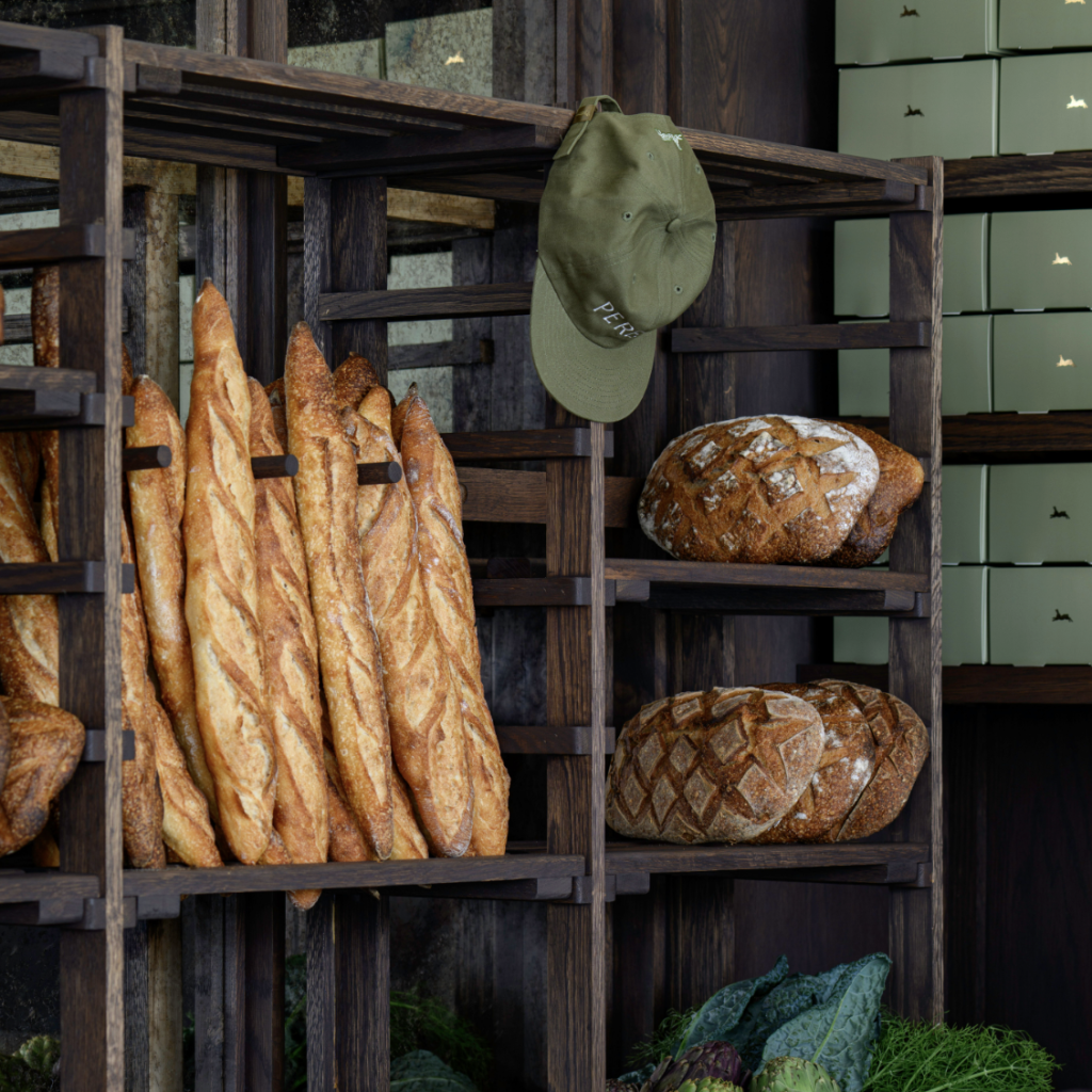 A rustic bakery display featuring artisanal breads and a green cap with 'PERE' embroidered on it, hanging from a wooden rack in Williamson County, Tennessee.