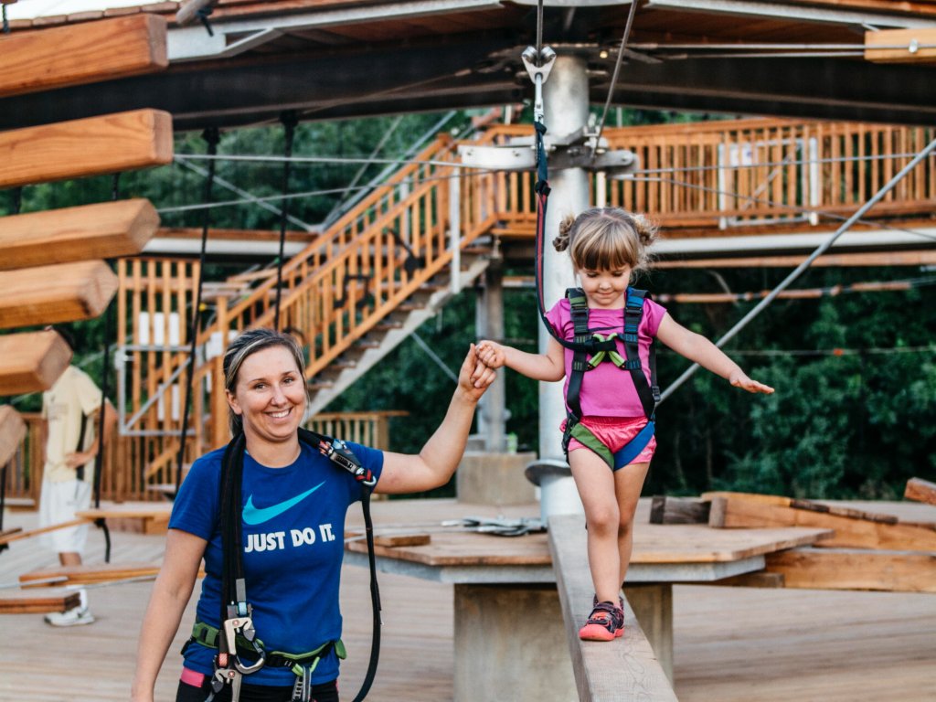 A smiling woman in a blue 'Just Do It' t-shirt helps a young girl in a pink shirt and harness walk across a suspended wooden beam at an adventure park in Williamson County, Tennessee.
