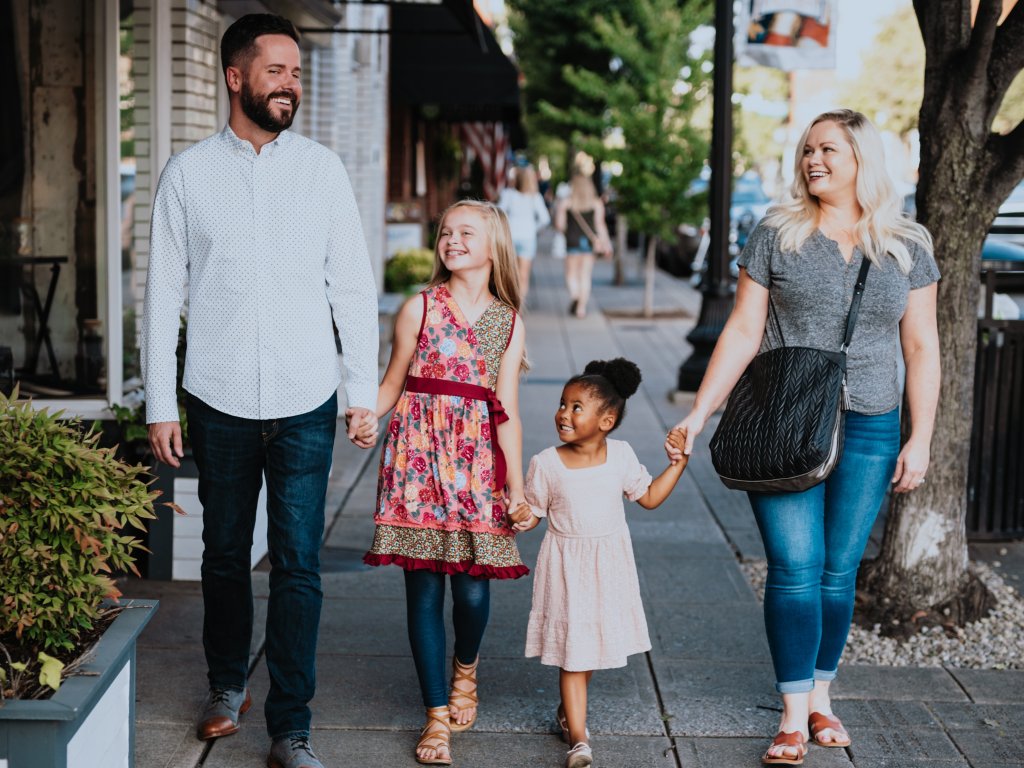 A family of four, including a man, woman, and two young girls, are walking hand in hand down a charming city sidewalk, smiling and enjoying each other's company in Williamson County, Tennessee.