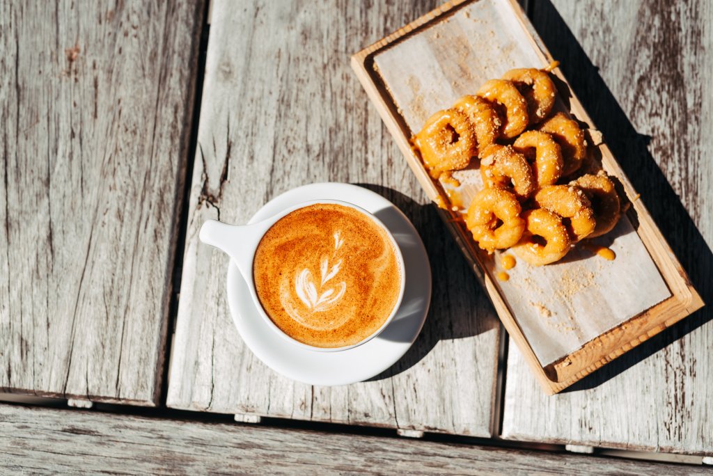 A steaming cup of latte art coffee sits beside a tray of golden-brown, sugar-coated churros on a rustic wooden table in Williamson County, Tennessee.