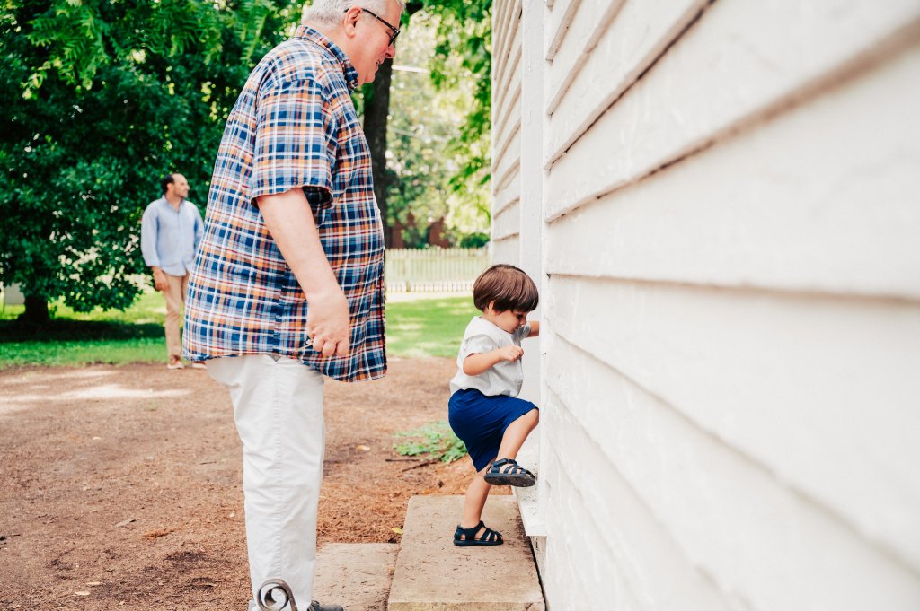 An elderly man in a plaid shirt assists a young boy in climbing the steps of a white building, while another man and a child look on from the background in Williamson County, Tennessee.