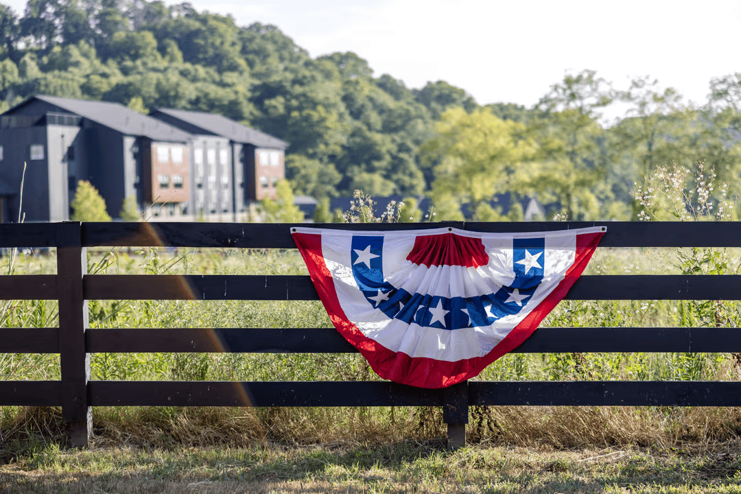 A patriotic bunting with red, white, and blue stars and stripes hangs from a black wooden fence, with a backdrop of lush green fields and modern buildings in Williamson County, Tennessee.