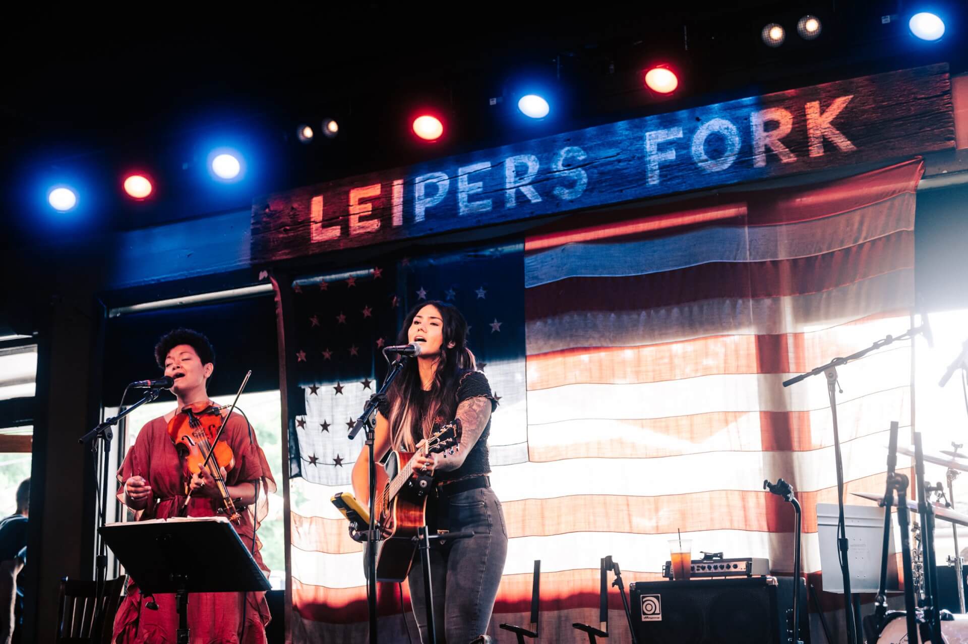 Two musicians perform on stage at Leipers Fork, one playing violin and the other singing and playing guitar, with an American flag backdrop and colorful stage lights in Williamson County, Tennessee.